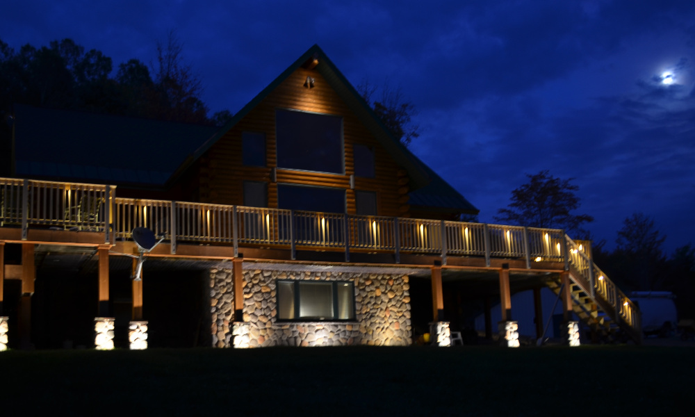 Stone and log A-frame cabin with outdoor lighting on pillars, railing, and roof peak